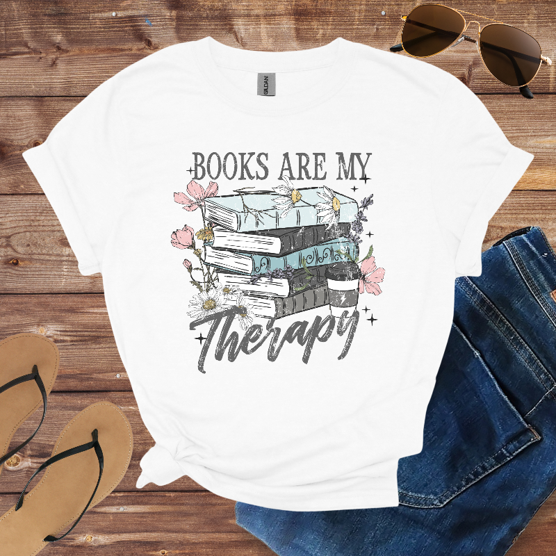 White t-shirt with 'Books are my Therapy' design on a wooden surface with sunglasses, jeans, and sandals.
