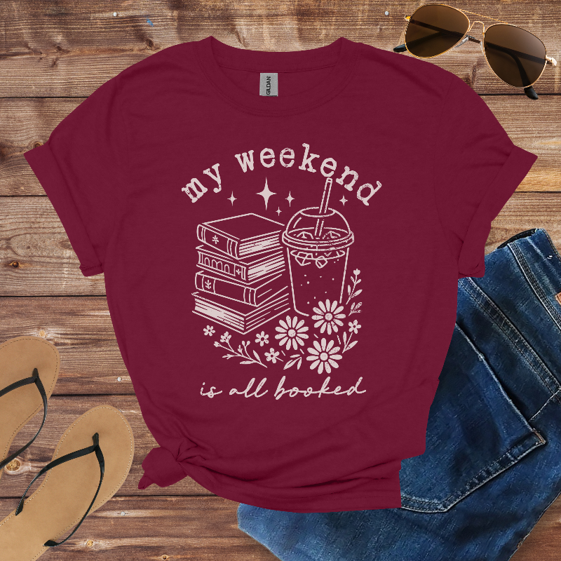 Maroon t-shirt with 'my weekend is all booked' design on a wooden surface with sunglasses, jeans, and flip-flops.