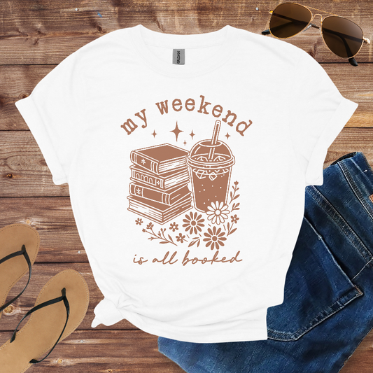 White t-shirt with 'my weekend is all booked' design on a wooden surface with sunglasses, jeans, and flip-flops.