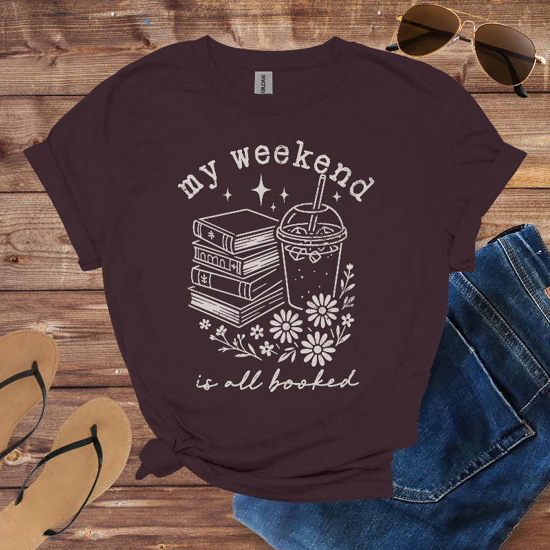 Brown t-shirt with 'my weekend is all booked' design on a wooden surface with sunglasses, jeans, and sandals.