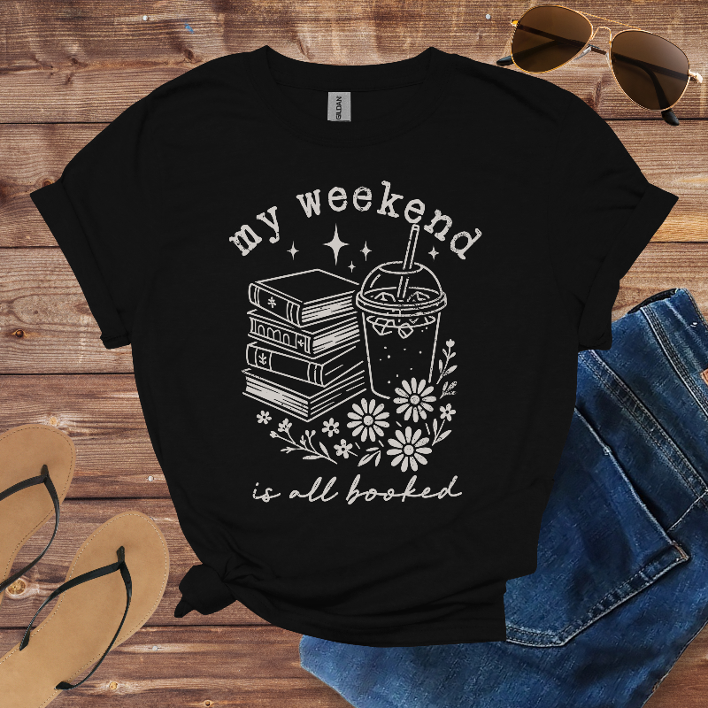 Black t-shirt with 'my weekend is all booked' design on a wooden surface with sunglasses, jeans, and flip-flops.
