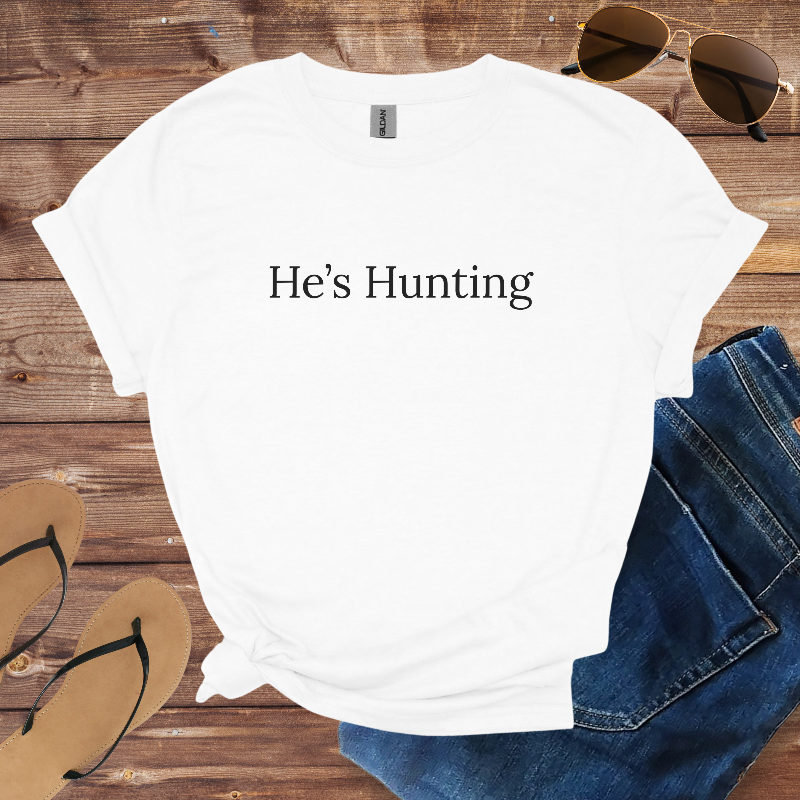 White t-shirt with 'He's Hunting' text on a wooden surface with sunglasses, jeans, and flip-flops.