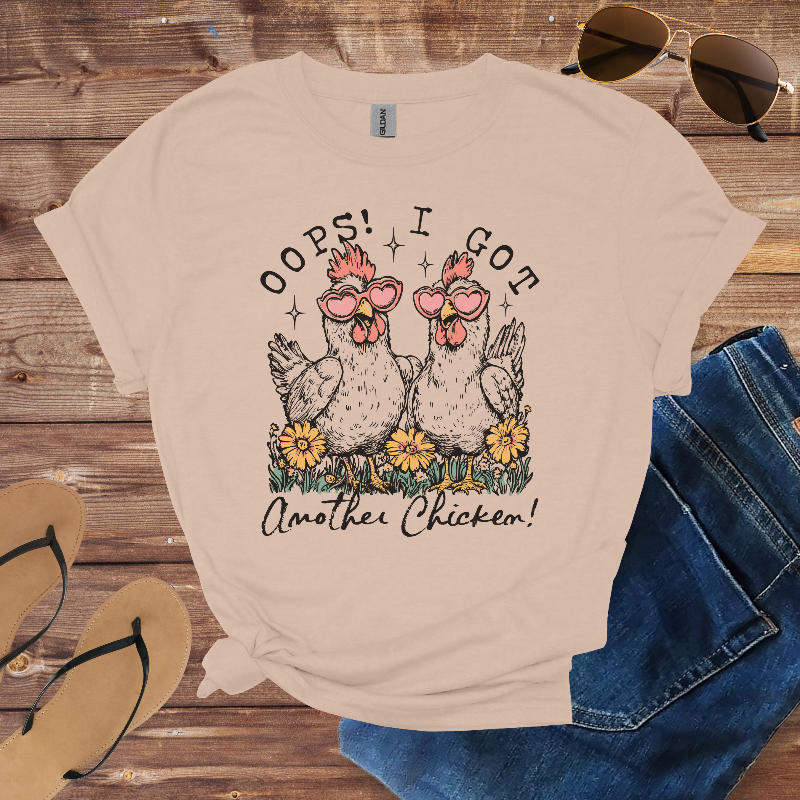 Beige t-shirt with chicken graphic and text on a wooden surface with sunglasses and jeans.